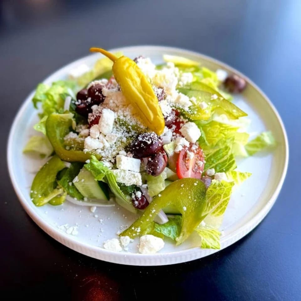 A vibrant Greek salad with lettuce, tomatoes, olives, feta, and a yellow pepperoncini on a white plate.