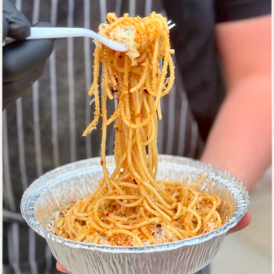 A person in a striped apron and black gloves holds a foil container of spaghetti with a forkful lifted.