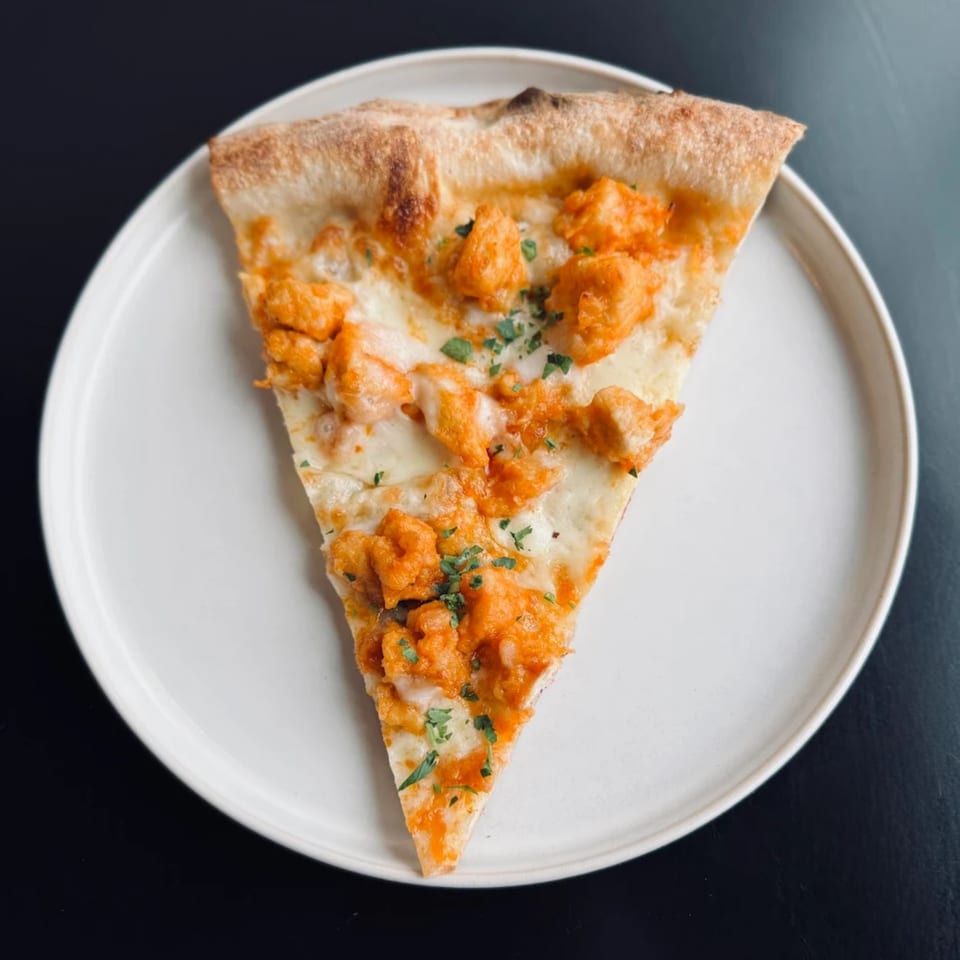 A single slice of pizza with white cheese, orange chicken pieces, and green herbs on a white plate.