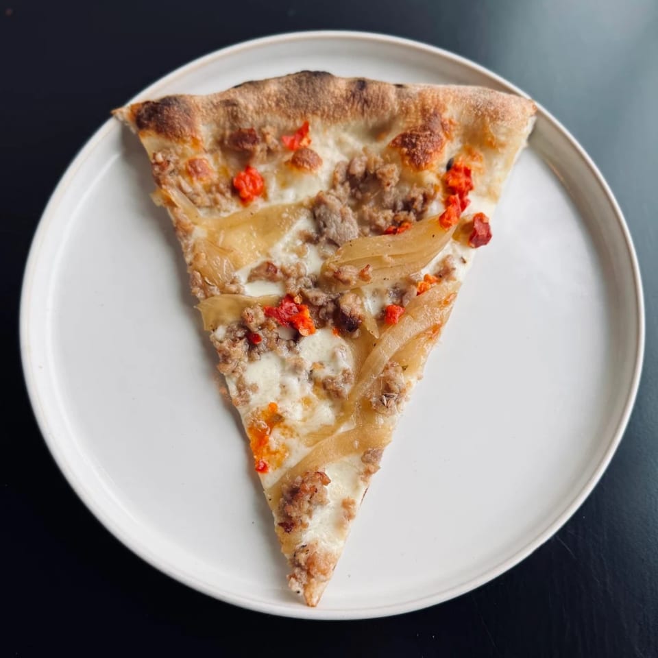 A single slice of white pizza with sausage, caramelized onions, and red peppers on a white plate.