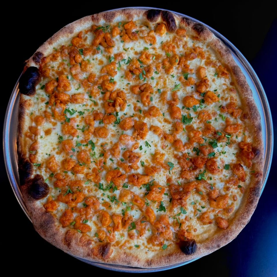 A whole round pizza with white cheese, orange chicken pieces, and green herbs on a metal tray.