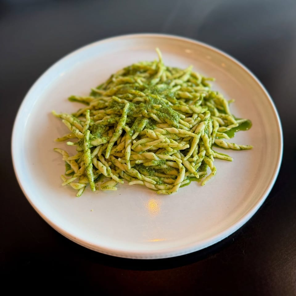 A white plate holds a serving of green pesto pasta, with twisted pasta noodles.