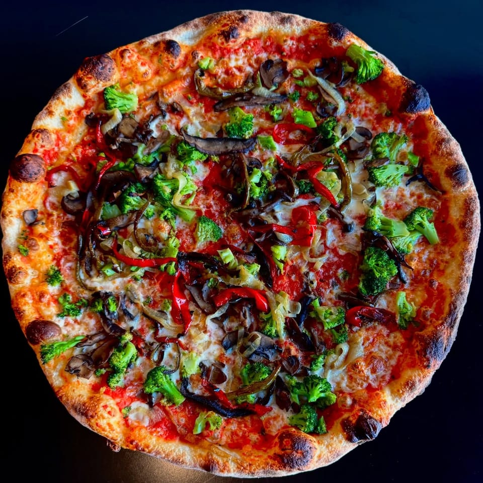 A vibrant vegetarian pizza with broccoli, mushrooms, onions, and red peppers on a dark surface.