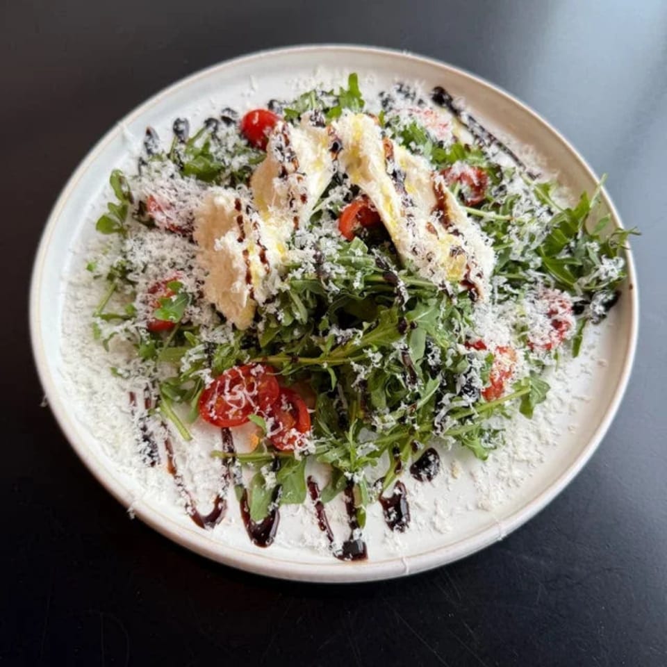 Arugula salad with cherry tomatoes, shaved parmesan, and balsamic glaze on a white plate.